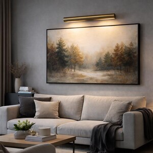 May include: A living room scene featuring a large, framed landscape painting above a light-colored sectional sofa. A brass-toned picture light illuminates the artwork. The room includes a side table with a lamp, and a coffee table.