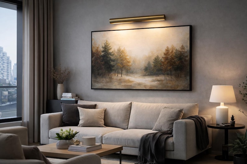 May include: A living room scene featuring a large, framed landscape painting above a light-colored sectional sofa. A brass-toned picture light illuminates the artwork. The room includes a side table with a lamp, and a coffee table.