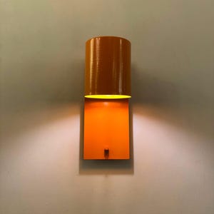 Op de afbeelding: Een feloranje wandlamp met een cilindrische kap en een rechthoekige basis. Het licht is aan en geeft een warm gloed.