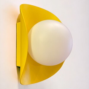 May include: A yellow wall sconce with a white globe light fixture. The sconce is shaped like a crescent moon.
