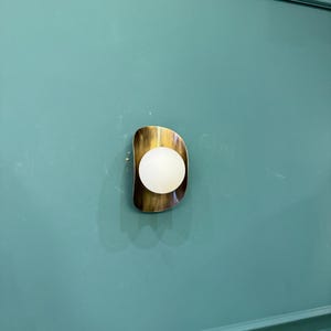 May include: A contemporary wall sconce with a brass-toned, freeform frame. It has a spherical, white light. The sconce is fixed to a teal-coloured wall.