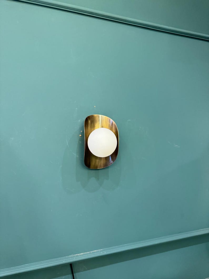 May include: A contemporary wall sconce with a brass-toned, freeform frame. It has a spherical, white light. The sconce is fixed to a teal-coloured wall.