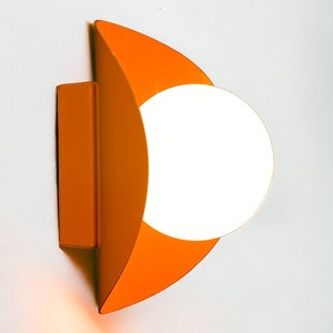 May include: Orange wall sconce with a white globe light fixture. The sconce is shaped like a crescent moon.