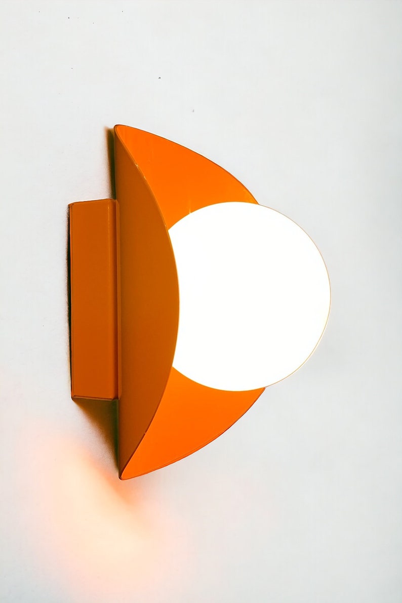 May include: Orange wall sconce with a white globe light fixture. The sconce is shaped like a crescent moon.