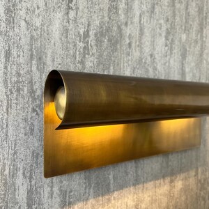 May include: A wall-mounted, brass-toned light fixture. The fixture has a rectangular base and a curved, cylindrical top that houses the light source. The light shines downward, creating a warm glow. The fixture is mounted on a textured gray wall.