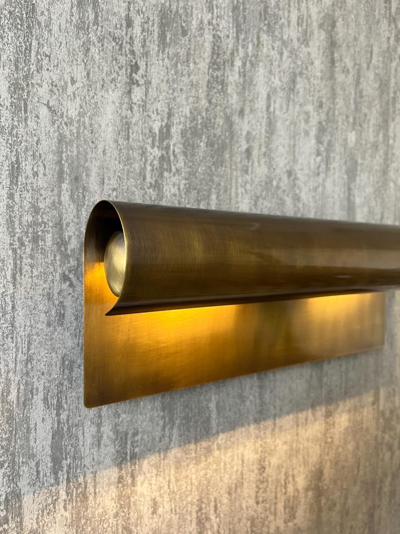 May include: A wall-mounted, brass-toned light fixture. The fixture has a rectangular base and a curved, cylindrical top that houses the light source. The light shines downward, creating a warm glow. The fixture is mounted on a textured gray wall.