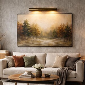 May include: A living room scene featuring a cream-colored sofa with several throw pillows in shades of brown and green. A landscape painting hangs above the sofa, and a round wooden coffee table sits in front. A lamp and decorative items complete the cozy setting.