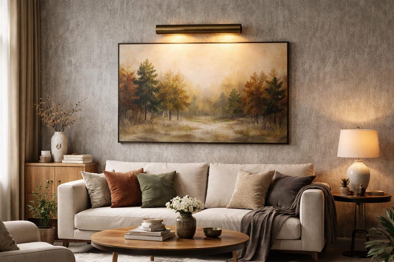 May include: A living room scene featuring a cream-colored sofa with several throw pillows in shades of brown and green. A landscape painting hangs above the sofa, and a round wooden coffee table sits in front. A lamp and decorative items complete the cozy setting.