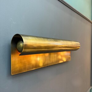 May include: A brass wall sconce with a rectangular backplate and a curved, cylindrical shade. The sconce has a warm, golden hue and is mounted on a gray wall. The light is on, emitting a soft glow.