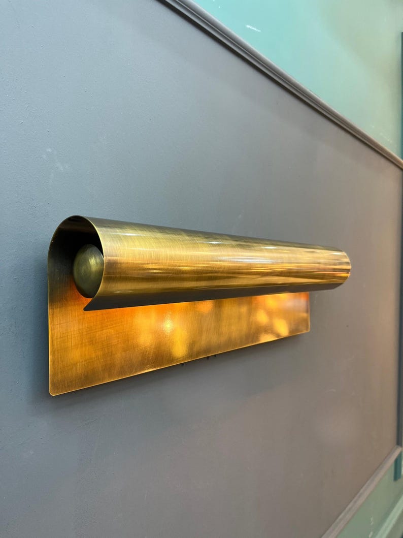 May include: A brass wall sconce with a rectangular backplate and a curved, cylindrical shade. The sconce has a warm, golden hue and is mounted on a gray wall. The light is on, emitting a soft glow.
