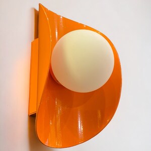 May include: Orange wall sconce with a white globe light fixture. The sconce is shaped like a curved leaf.