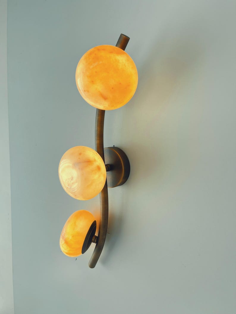 May include: A wall sconce with three glowing, spherical lights. The lights are a warm yellow-orange color, and the fixture has a curved, dark brown metal stem. The sconce is mounted on a wall, providing ambient lighting.