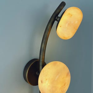 May include: A modern wall sconce with a curved, dark metal arm and two oval-shaped, glowing light fixtures. The light fixtures have a warm, yellow-orange color, and the sconce is mounted on a light blue wall.
