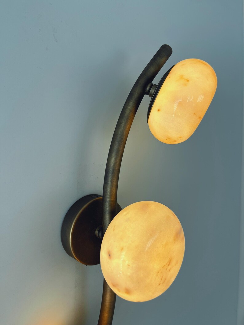 May include: A modern wall sconce with a curved, dark metal arm and two oval-shaped, glowing light fixtures. The light fixtures have a warm, yellow-orange color, and the sconce is mounted on a light blue wall.