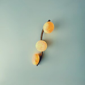 May include: A modern wall sconce featuring three spherical, glowing orange lights. The lights are arranged vertically along a curved, dark brown metal arm. The sconce is mounted on a light blue wall.