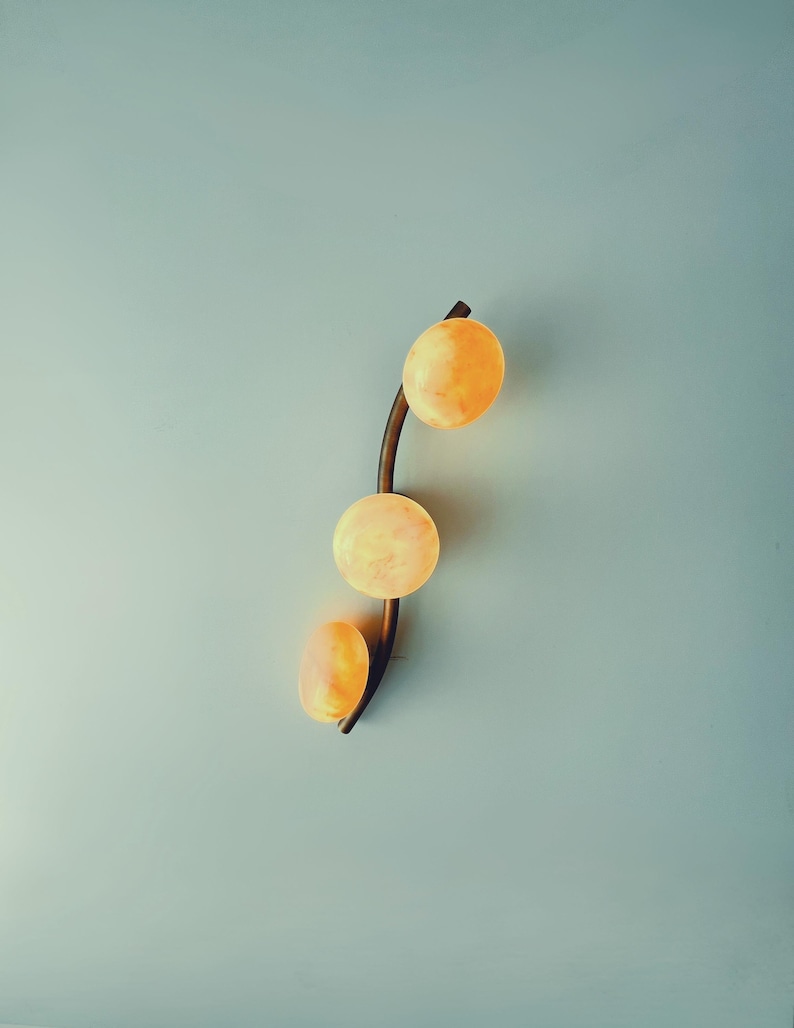 May include: A modern wall sconce featuring three spherical, glowing orange lights. The lights are arranged vertically along a curved, dark brown metal arm. The sconce is mounted on a light blue wall.