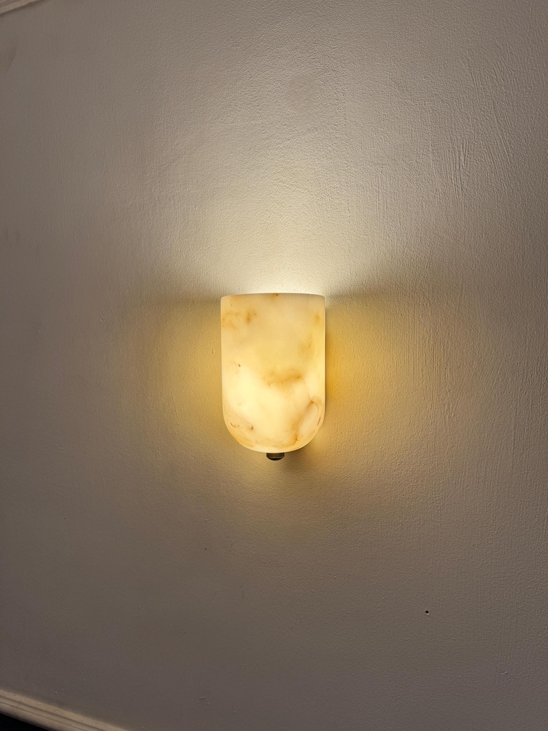 May include: A wall-mounted sconce with a warm, glowing light. The fixture is made of a translucent, beige-colored material with subtle veining. The sconce has a rounded top and a small, dark-colored base.