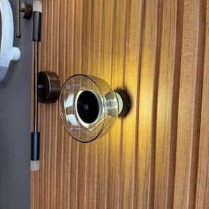 May include: A clear glass globe wall sconce with a black interior, mounted on a wooden panel. The sconce has a circular design and is illuminated. Additional lighting fixtures are visible in the background.