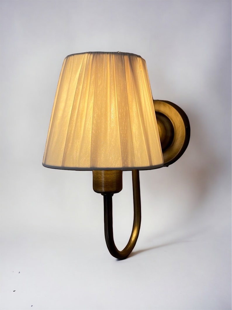 May include: A gold wall sconce with a pleated fabric lampshade. The lampshade is a light beige colour.