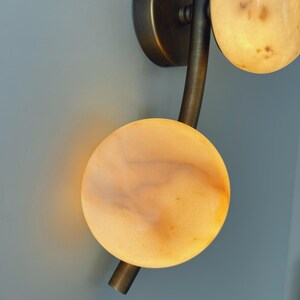 May include: A wall sconce with a bronze finish and three circular, glowing alabaster shades. The fixture has a curved arm and a round base, mounted on a light blue wall. The alabaster shades have a warm, amber glow.