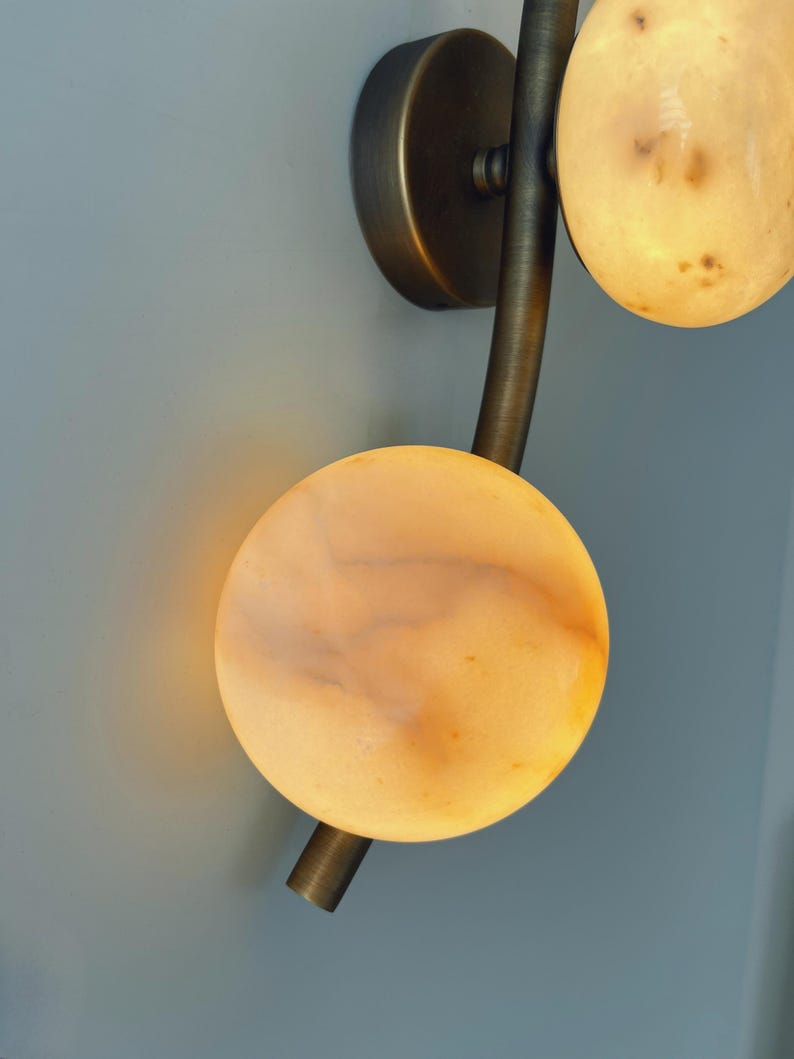 May include: A wall sconce with a bronze finish and three circular, glowing alabaster shades. The fixture has a curved arm and a round base, mounted on a light blue wall. The alabaster shades have a warm, amber glow.