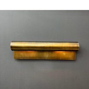May include: A brass-colored wall sconce with a rectangular base and a cylindrical top. The sconce emits a warm, inviting light. The fixture is mounted on a gray wall, creating a contrast with the metallic finish. This modern lighting fixture adds a touch of elegance.