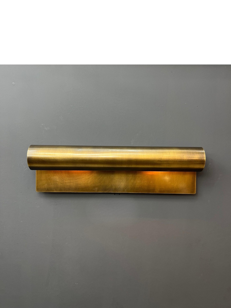May include: A brass-colored wall sconce with a rectangular base and a cylindrical top. The sconce emits a warm, inviting light. The fixture is mounted on a gray wall, creating a contrast with the metallic finish. This modern lighting fixture adds a touch of elegance.