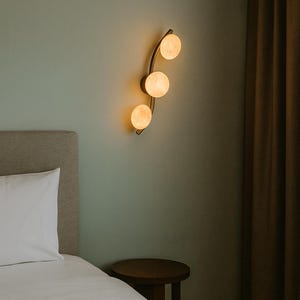 May include: A modern wall sconce with three spherical, glowing lights. The fixture has a curved, brass-colored arm and is mounted on a pale green wall. The scene includes a bed with a white pillow and a small wooden side table.