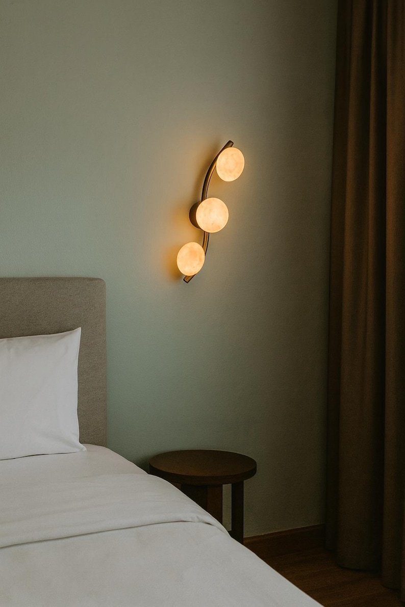 May include: A modern wall sconce with three spherical, glowing lights. The fixture has a curved, brass-colored arm and is mounted on a pale green wall. The scene includes a bed with a white pillow and a small wooden side table.