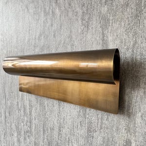 May include: A modern, wall-mounted light fixture with a curved, cylindrical shade and a rectangular base. The fixture is made of a warm, metallic brass material and is mounted on a textured gray wall.