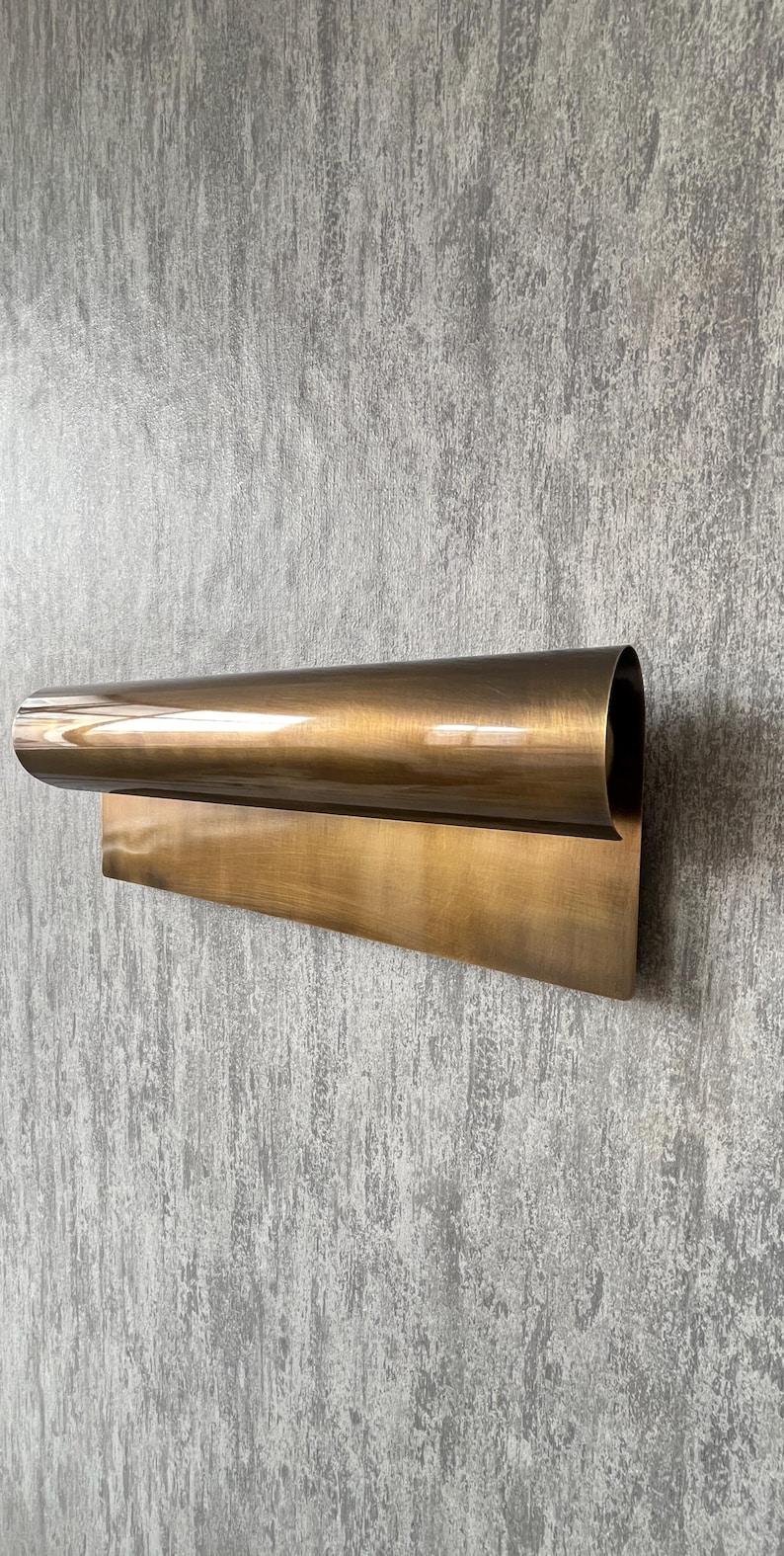 May include: A modern, wall-mounted light fixture with a curved, cylindrical shade and a rectangular base. The fixture is made of a warm, metallic brass material and is mounted on a textured gray wall.