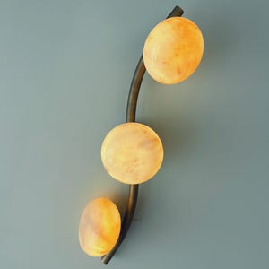 May include: A modern wall sconce featuring three glowing, oval-shaped lights in shades of yellow and orange. The lights are arranged along a curved, dark metal stem against a muted blue-gray background. The fixture is designed for interior lighting.