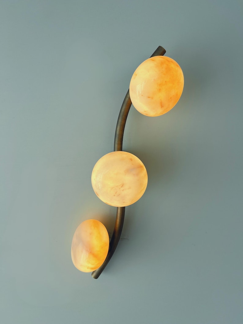 May include: A modern wall sconce featuring three glowing, oval-shaped lights in shades of yellow and orange. The lights are arranged along a curved, dark metal stem against a muted blue-gray background. The fixture is designed for interior lighting.