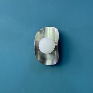 May include: A contemporary wall light featuring a brushed metal finish and a spherical white bulb. The light has a curved, elongated shape and is mounted on a teal blue wall. The light fixture provides a soft, ambient glow.