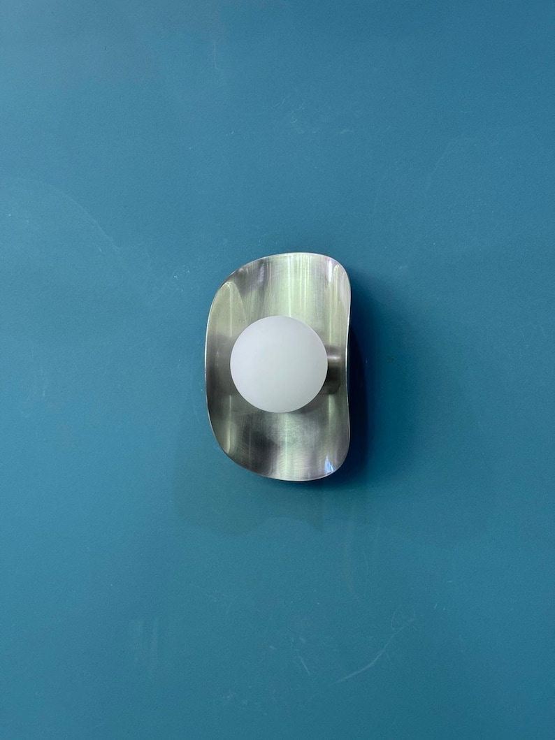May include: A contemporary wall light featuring a brushed metal finish and a spherical white bulb. The light has a curved, elongated shape and is mounted on a teal blue wall. The light fixture provides a soft, ambient glow.