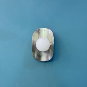 May include: A contemporary wall light with a brushed metal finish. The light has a rounded, elongated shape with a white globe bulb in the centre. The light is mounted on a blue wall, offering a minimalist and modern aesthetic.