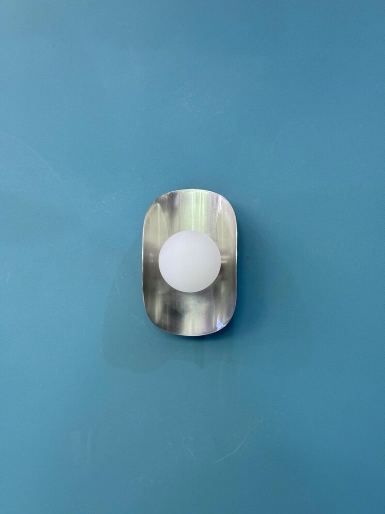May include: A contemporary wall light with a brushed metal finish. The light has a rounded, elongated shape with a white globe bulb in the centre. The light is mounted on a blue wall, offering a minimalist and modern aesthetic.