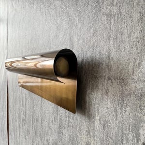 May include: A modern, wall-mounted sconce with a curved, metallic design. The sconce has a warm, brass-like finish and a cylindrical opening that likely houses a light source. The fixture is mounted on a textured, gray wall.