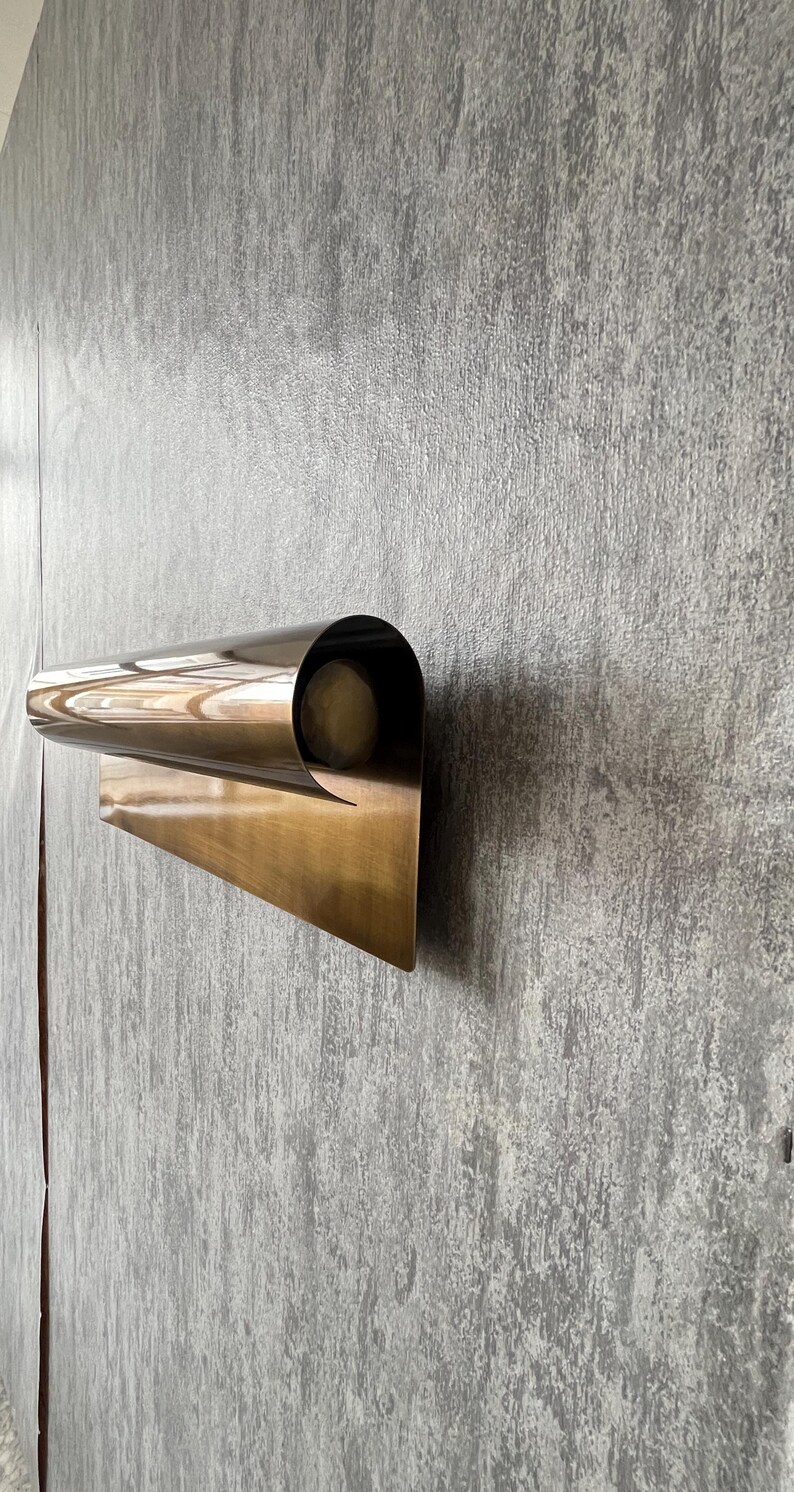 May include: A modern, wall-mounted sconce with a curved, metallic design. The sconce has a warm, brass-like finish and a cylindrical opening that likely houses a light source. The fixture is mounted on a textured, gray wall.