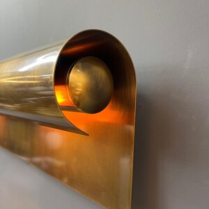 May include: A brass wall sconce with a unique, curved design. The sconce features a cylindrical element that curls around a spherical accent, with a triangular base. The interior glows with warm light, creating a striking visual effect.