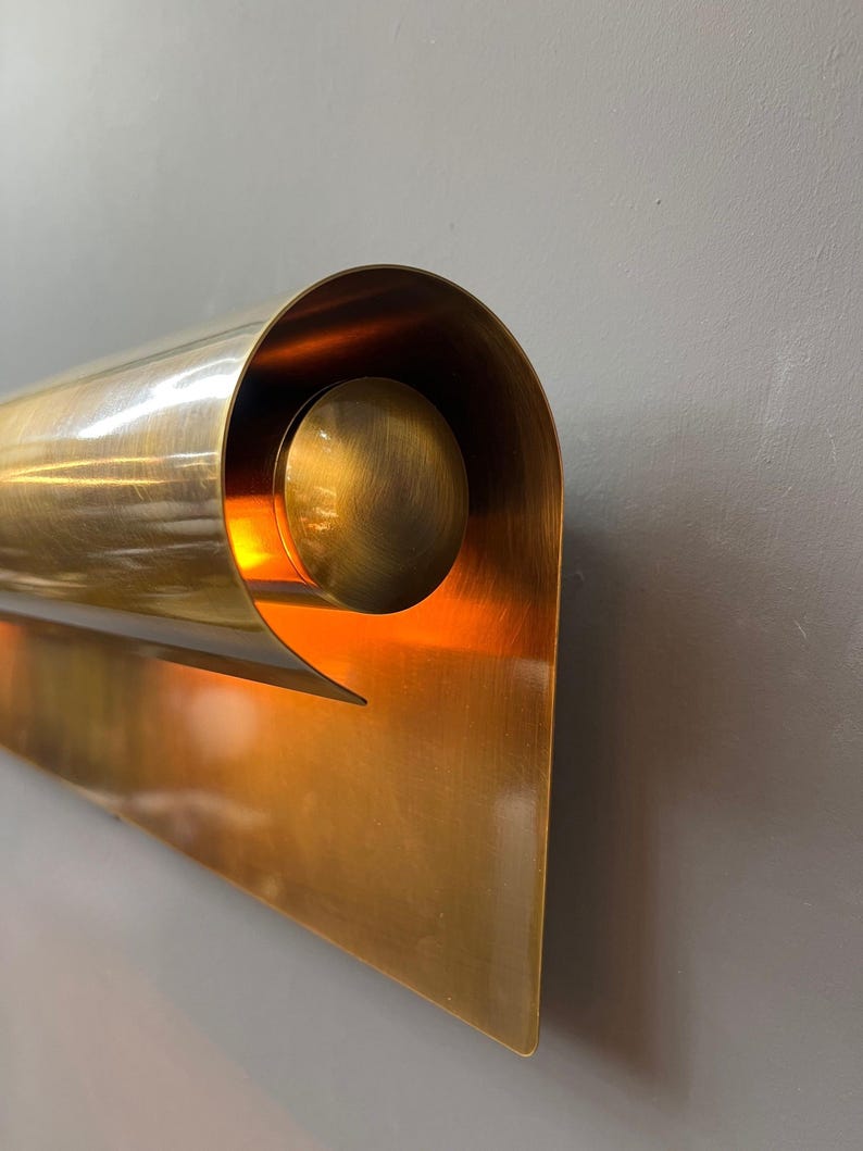 May include: A brass wall sconce with a unique, curved design. The sconce features a cylindrical element that curls around a spherical accent, with a triangular base. The interior glows with warm light, creating a striking visual effect.