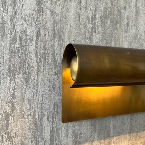 May include: A close-up of a brass wall sconce with a curved top and rectangular base. The sconce is mounted on a textured gray wall. The light from the sconce illuminates the wall.