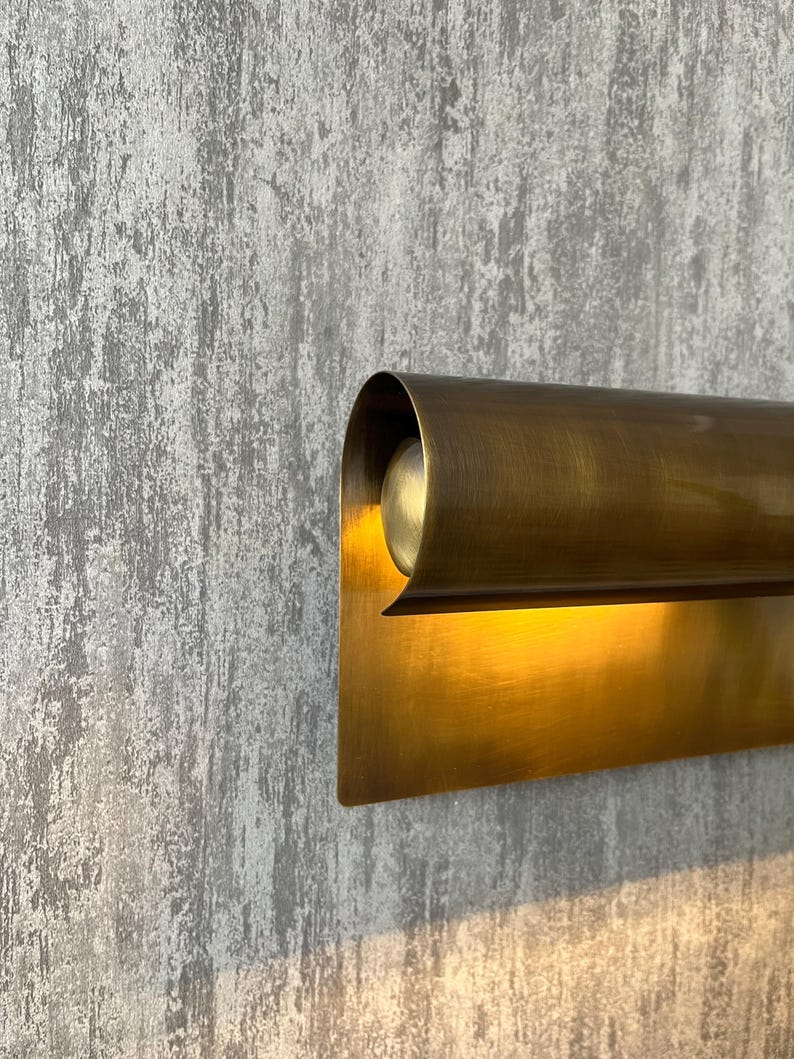 May include: A close-up of a brass wall sconce with a curved top and rectangular base. The sconce is mounted on a textured gray wall. The light from the sconce illuminates the wall.