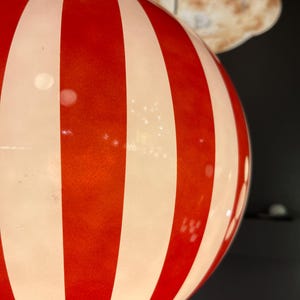May include: A round, red and white striped light fixture. The fixture has alternating vertical stripes of red and white. The light fixture is hanging from the ceiling. The background is dark.