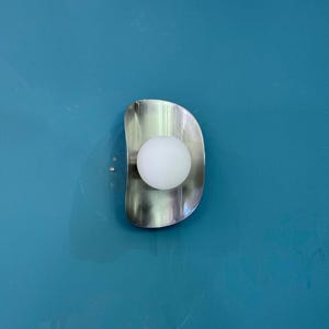 May include: A contemporary wall light with a brushed metal finish and a spherical white bulb. The light fitting has a curved, organic shape and is mounted on a teal wall. The light fixture provides a soft, ambient glow.