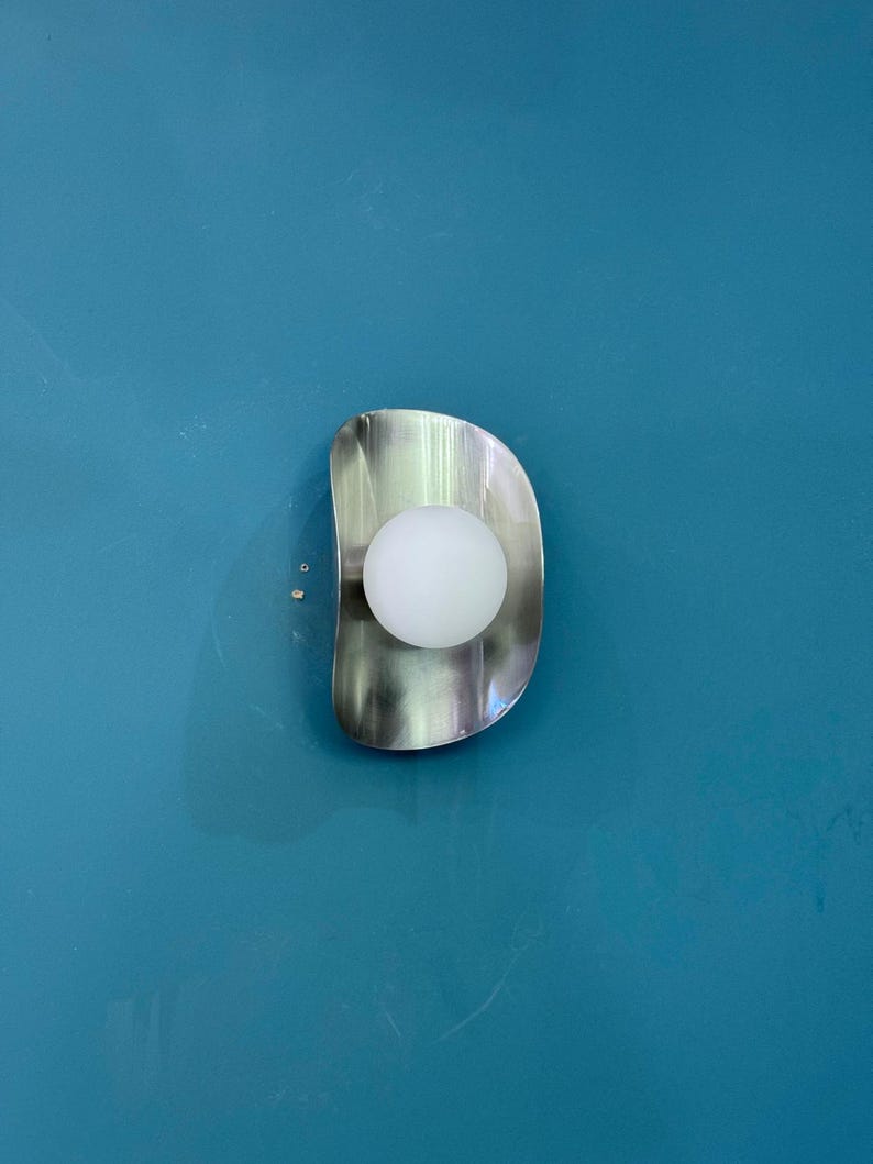May include: A contemporary wall light with a brushed metal finish and a spherical white bulb. The light fitting has a curved, organic shape and is mounted on a teal wall. The light fixture provides a soft, ambient glow.