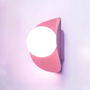 May include: A pink wall-mounted light fixture with a round, white light bulb. The light fixture is shaped like a crescent moon.