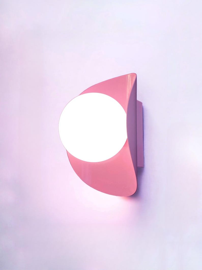 May include: A pink wall-mounted light fixture with a round, white light bulb. The light fixture is shaped like a crescent moon.