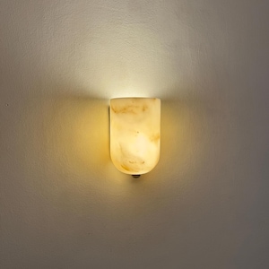 May include: A wall-mounted light fixture with a rounded top and a warm, glowing light. The fixture is made of a translucent, marbled material, likely stone or alabaster, and casts a soft, inviting illumination on the wall.