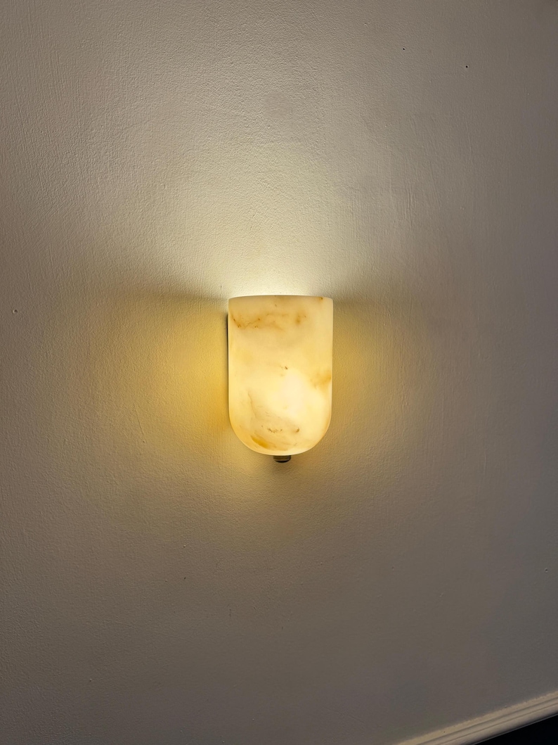May include: A wall-mounted light fixture with a rounded top and a warm, glowing light. The fixture is made of a translucent, marbled material, likely stone or alabaster, and casts a soft, inviting illumination on the wall.